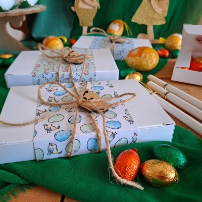 Caixas brancas decoradas com papel estampado de ovos da Páscoa, amarradas com cordão de juta e ornamento de coelho, com ovos de chocolate coloridos à volta