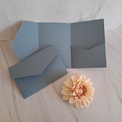 Convite aberto azul claro em papel texturizado sobre superfície tipo mármore com flor creme próxima