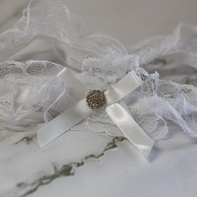 Liga nupcial branca em renda com laço de cetim e botão prateado sobre mármore
