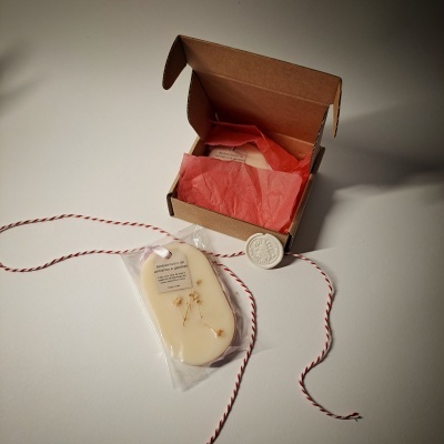Sabonete artesanal branco com flores secas, cordão vermelho e branco, embalagem em plástico transparente, caixa castanha aberta com papel de seda vermelho e selo branco.