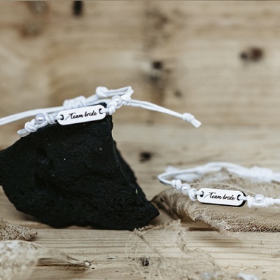 Braceletes brancos com placa 'Team bride' gravada, sobre madeira e pedra preta