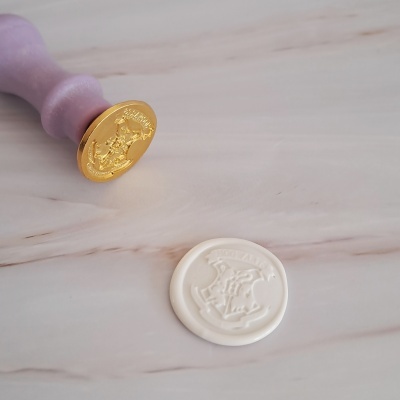Carimbo de lacre dourado com cabo roxo e selo de cera branca com escudo Hogwarts