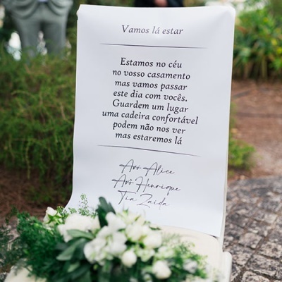 Papel branco com texto em português preso a uma cadeira branca decorada com flores brancas e verdes em ambiente exterior