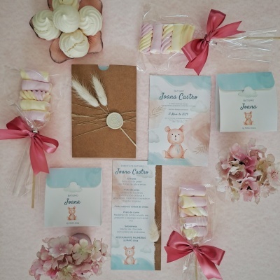 Artigos para evento com tema ursinho em tons rosa e azul, com convite, embalagens, doces e flores sobre fundo rosa.