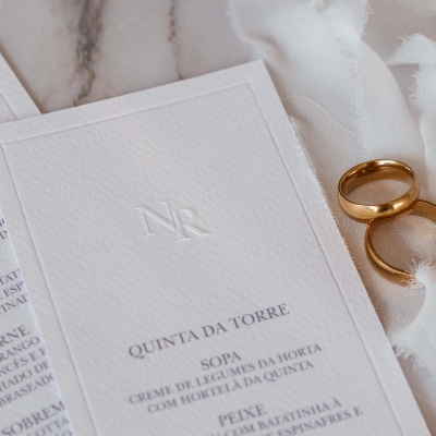 Convite ou menu branco texturizado com letras em relevo e texto impresso, ao lado dois anéis dourados sobre tecido branco