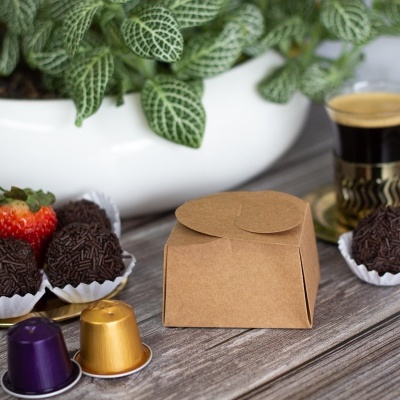 Caixa de papel castanho com brigadeiros, morango, cápsulas de café, chávena de café e planta verde