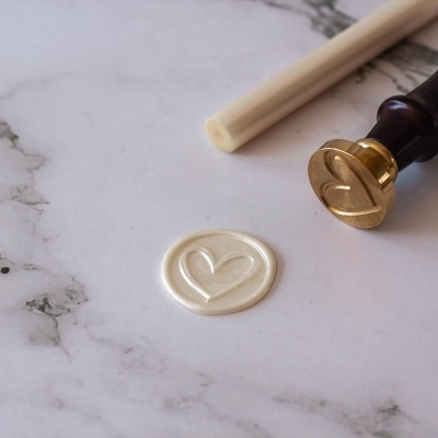 Selo de cera bege com coração, carimbo dourado com cabo de madeira e bastão de cera sobre mármore claro