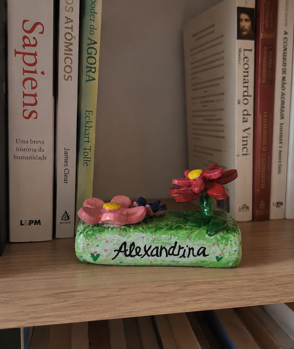 Jardim | Peça Personalizada com Nome Suporte decorativo verde com flores em cerâmica e nome Alexandrina, numa prateleira com livros