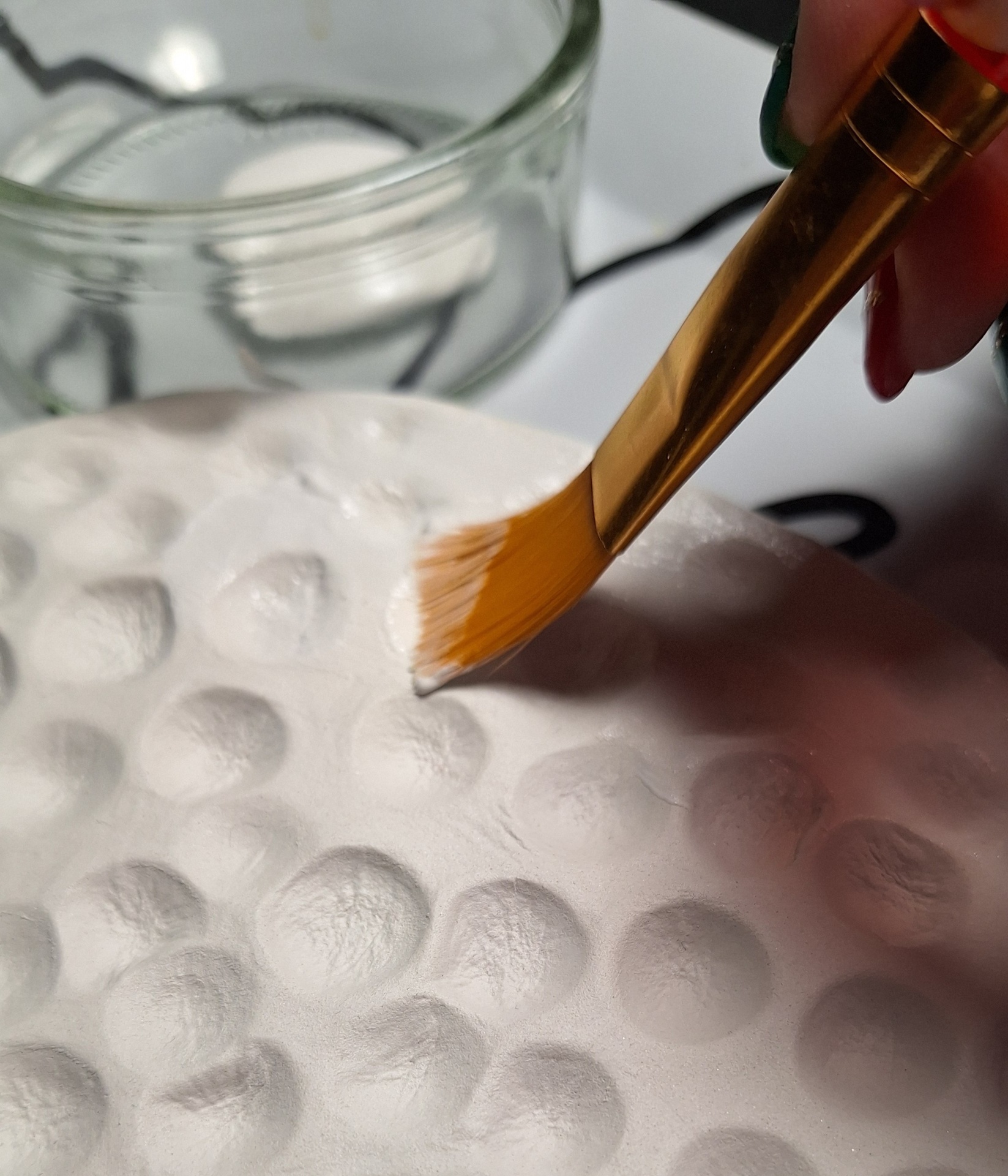 Pincel dourado aplicando produto branco sobre superfície texturizada com padrão de bolhas.