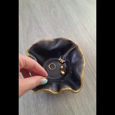 Taça negra com textura e bordas douradas, mãos segurando, anel e colar dourados no interior.