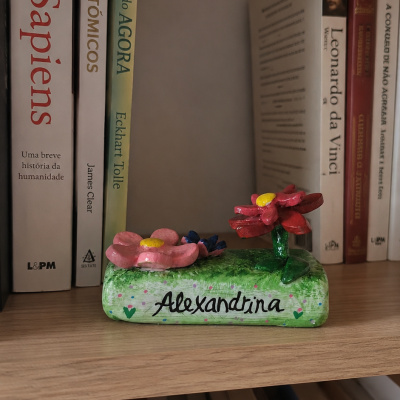 Suporte decorativo verde com flores em cerâmica e nome Alexandrina, numa prateleira com livros