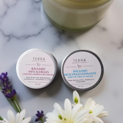 Dois bálsamos pequenos em latas de metal da marca TERRA sobre superfície branca marmorizada com flores brancas e lilás e um frasco grande ao fundo.