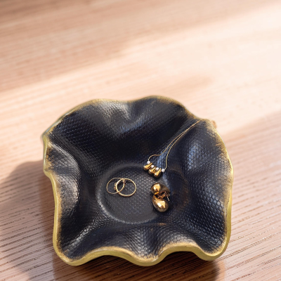 Taça decorativa preta com textura de pele de serpente e bordas douradas com joias douradas dentro, sobre madeira clara.