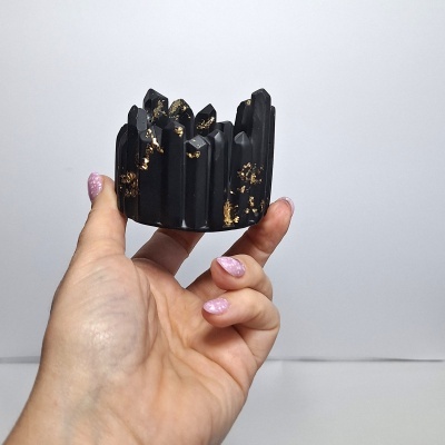 Escultura preta com detalhes dourados em formato de cristais segurada por mão com unhas rosa glitter