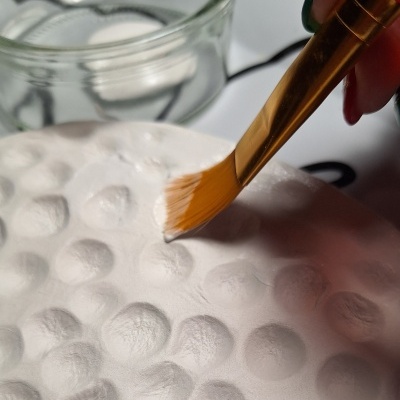 Pincel dourado aplicando produto branco sobre superfície texturizada com padrão de bolhas.