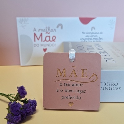 Etiqueta rosa com texto dourado sobre o amor de mãe, flores roxas, livro e placa de fundo