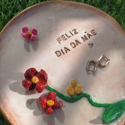 Prato cerâmico com flores esmaltadas, texto FELIZ DIA DA MÃE e brincos prateados sobre relva verde