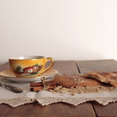 Conjunto de taça e prato amarelos com ilustração, faca decorada, pão rústico e toalha branca numa mesa de madeira.