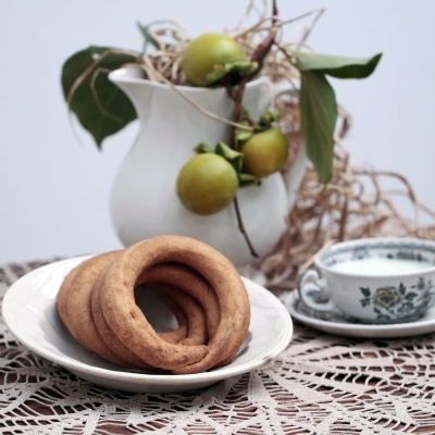 Biscoitos redondos dourados em prato branco com chávena azul floral, toalha de renda e vaso branco com frutas e folhas.