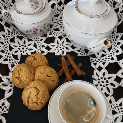 Conjunto de chá em porcelana branca com flores rosas, café numa chávena, bolachas e paus de canela numa base preta sobre renda branca.