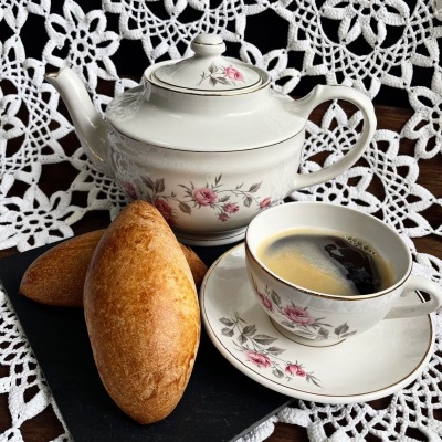 Conjunto de chá em porcelana com flores rosas, chávena com café, bule e dois pães dourados sobre toalha rendada branca