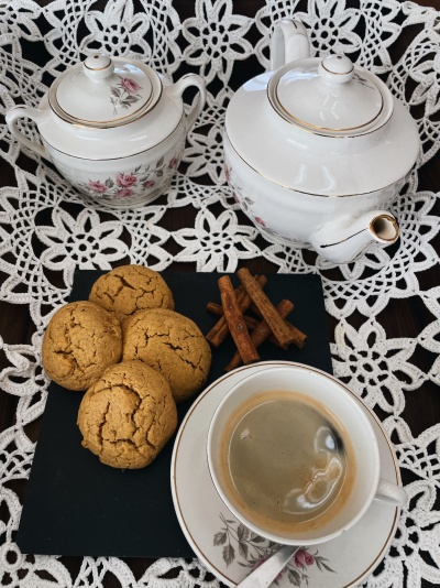 Conjunto de chá em porcelana branca com flores rosas, café numa chávena, bolachas e paus de canela numa base preta sobre renda branca.