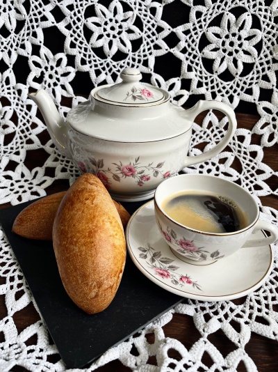 Conjunto de chá em porcelana com flores rosas, chávena com café, bule e dois pães dourados sobre toalha rendada branca