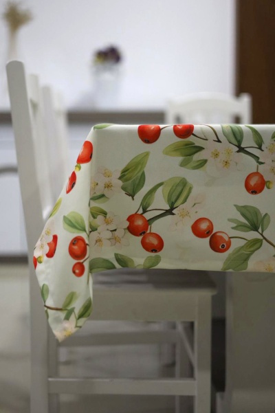 Toalha de mesa creme com padrão de cerejas vermelhas e folhas verdes, sobre mesa com cadeiras brancas.