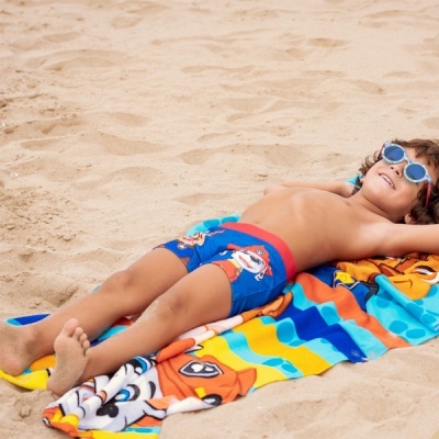 Criança deitada na praia com toalha colorida e calções azuis de desenho animado
