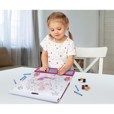 Criança a pintar desenho de sereia num caderno de colorir grande com lápis e tintas numa mesa branca