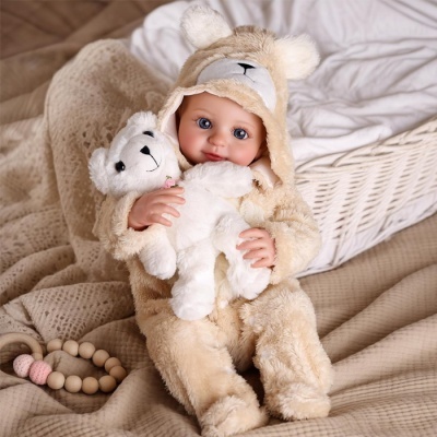 Boneca bebé com fato de urso bege segurando urso de peluche branco sentado num cenário com tecidos e cesta de vime.