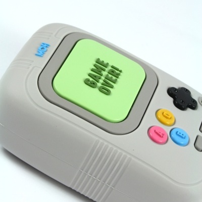 Comando de videojogo cinzento com botão verde 'GAME OVER!' e botões coloridos