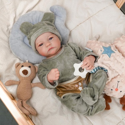 Boneco bebé realista com fato de veludo verde, deitado numa cama com almofada cinza e brinquedos de peluche castanho e rosa