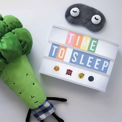 Brinquedo de peluche verde, máscara de dormir cinzenta e caixa luminosa com texto e emojis