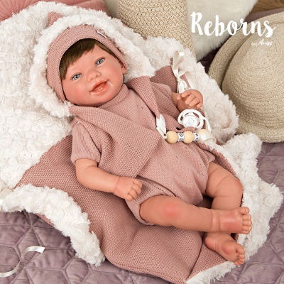 Boneca reborn com roupa rosa de malha deitada num autocolchão branco, com almofadas castanhas e bege ao fundo