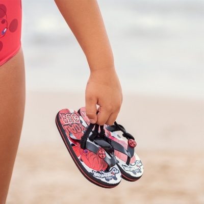 Sandálias de criança vermelhas e pretas com personagens, na mão de uma criança, fundo praia