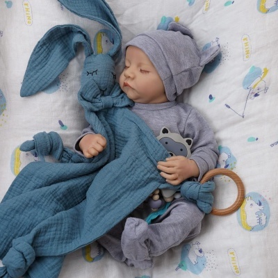 Boneca bebé com pijama cinza a segurar brinquedo de pano azul sobre lençol branco com unicórnios