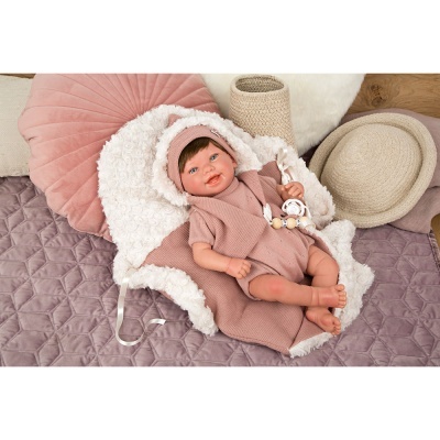 Boneca realista vestida em rosa sobre manta branca felpuda num ambiente com almofada rosa e objetos neutros