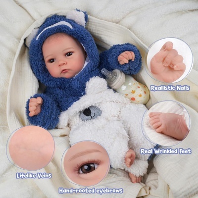 Boneca bebé realista de fato peludo azul e branco com capuz, segurando biberão com padrão de gatinhos.