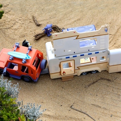brinquedo de carro vermelho com prancha de surf azul e caravana branca na areia