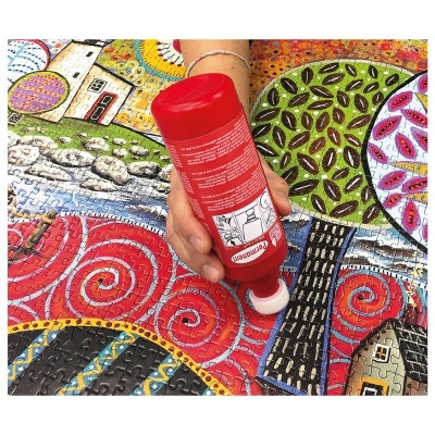 Mão aplicando cola vermelha num puzzle colorido com padrões abstratos e casas desenhadas.