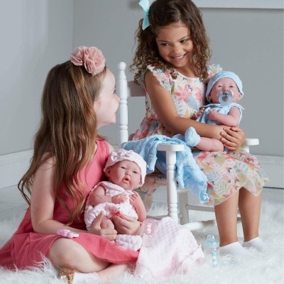 Meninas segurando bonecas realistas com vestidos coloridos, sentadas num tapete branco com cadeira ao fundo