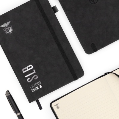 Cadernos pretos com cantos arredondados e elástico, um aberto mostrando página com linhas e símbolo, caneta preta ao lado.