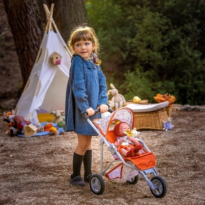 Criança com carrinho de bonecas vermelho e branco ao ar livre junto a tenda branca e brinquedos