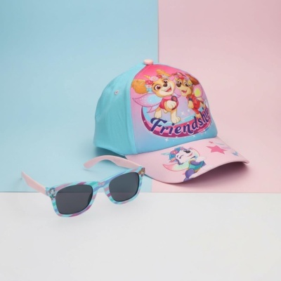 Boné infantil azul e rosa com desenhos de cães e óculos de sol complementares numa superfície branca com fundo azul e rosa.