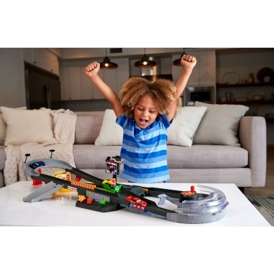 Menino com camisa azul brinca com pista de carrinhos em mesa branca, sofá e cozinha ao fundo