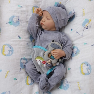 Bebé vestido com fato cinzento e chapéu com orelhas, a segurar brinquedo de raposa, deitado em manta branca com padrões