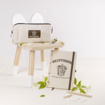 Estojo e caderno de tecido bege com tema Gryffindor, sobre banco de madeira claro com pernas brancas, decorados com folhas e flores brancas.