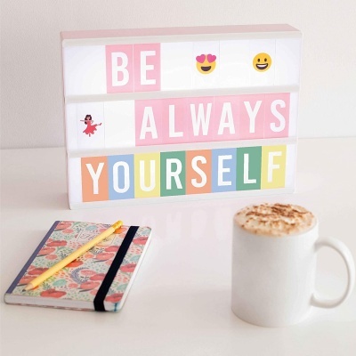 Caixa de luz com a frase BE ALWAYS YOURSELF, caderno floral, caneta amarela e caneca de café com espuma