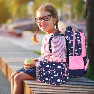Mochila e lancheira azul com padrões de nuvens e arco-íris junto a criança sentada num banco de madeira ao ar livre